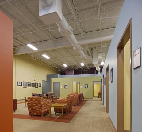 A modern lounge area featuring patterned chairs, tables, framed photos on pastel-colored walls, and an exposed ceiling with visible ducts. Several doors are aligned along the right side of the hallway.
