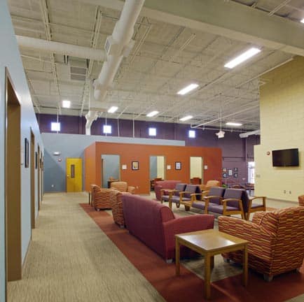 A spacious lounge features colorful chairs and couches, tables, and vibrantly painted walls. Large ceiling pipes and lights hang above, with several doors lining the side hallway.