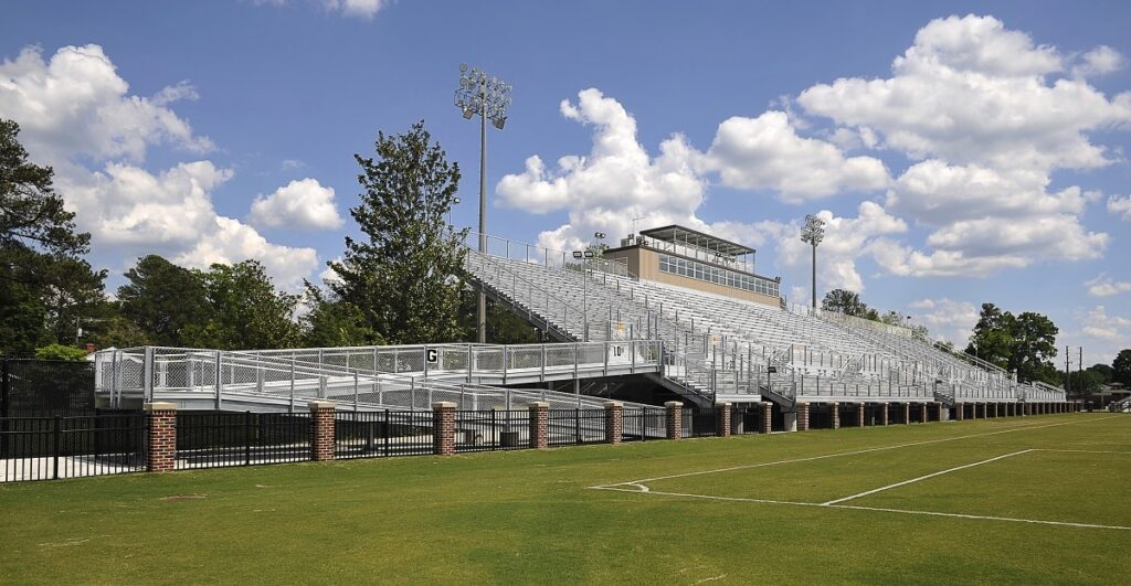Expansive empty metal bleachers stand beside a green sports field, surrounded by light poles and trees under a partly cloudy blue sky.