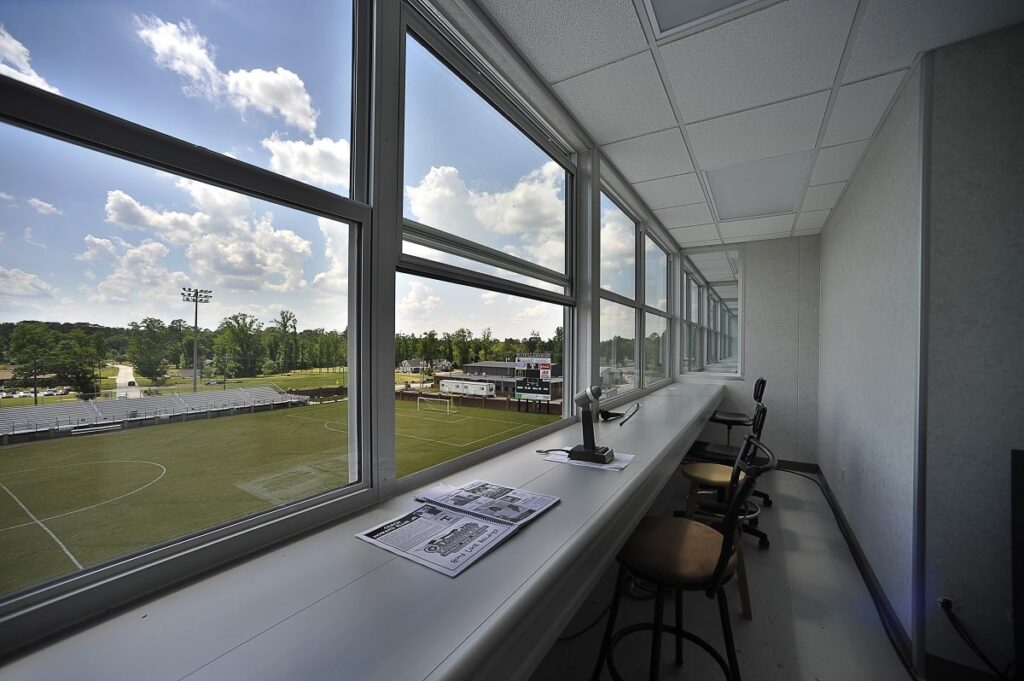 A scene from a press box with large windows providing a clear view of a soccer field, featuring several chairs and a desk cluttered with papers and a microphone, all illuminated by daylight under a bright sky.