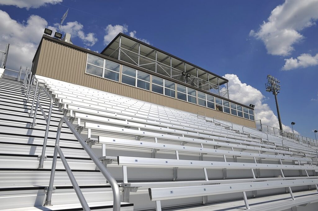 Rows of empty metal bleachers in an outdoor stadium, topped by a large press box, sit beneath a blue sky dotted with scattered clouds.