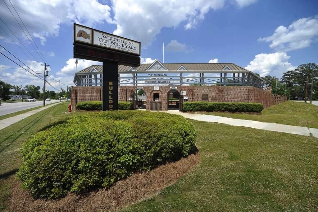 Entrance to a brick sports stadium with a sign reading Welcome to the Brickyard and Bulldog. Bushes and a sidewalk are in the foreground; a road runs along the left side. Blue sky with scattered clouds above.