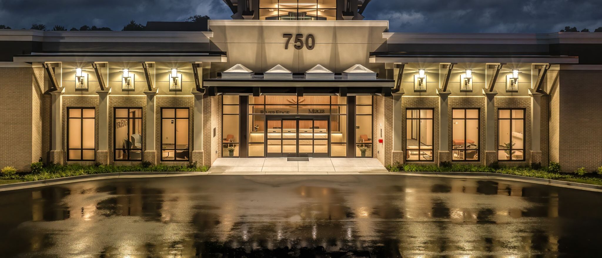 A brightly lit modern building with 750 above the entrance reflects on wet pavement at dusk beneath a dramatic, cloudy sky.