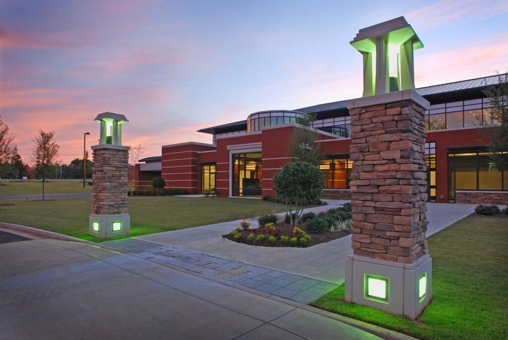 Modern brick building at sunset with large windows, a well-maintained lawn, and two stone pillars topped with green-lit lanterns framing the entrance. The pink and blue sky adds to the serene atmosphere.
