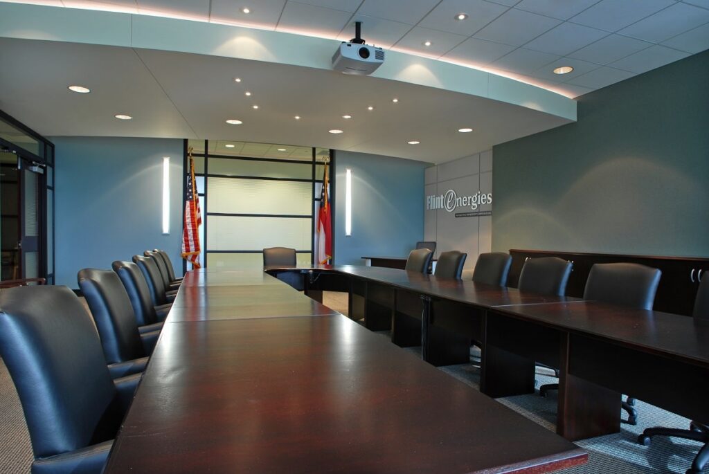 Modern conference room with a long, dark wood table, black leather chairs, flags, and a projector on the ceiling. The wall displays the Flint Energies logo. Room is empty and well-lit.