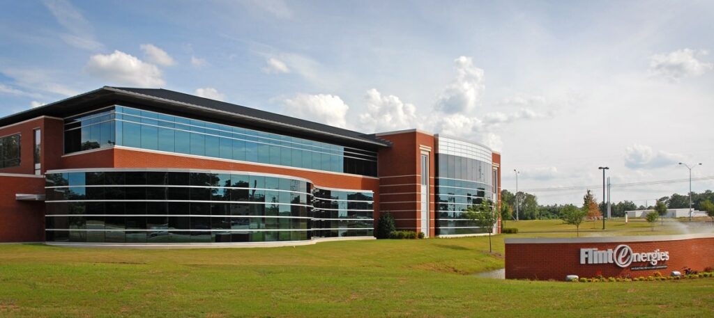 A modern brick office building with large glass windows sits behind a sign reading Flint Energies, surrounded by grass and trees under a partly cloudy sky.