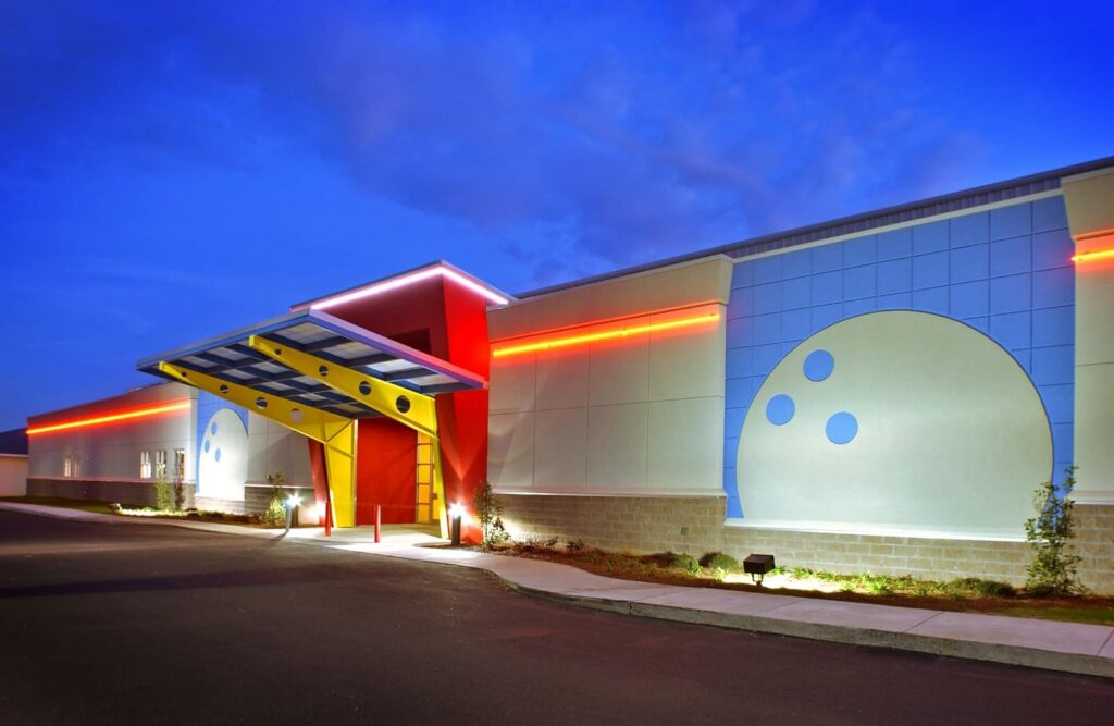 Modern bowling alley exterior at dusk, featuring large bowling ball graphics on the facade, a bright red entrance canopy, and colorful neon accent lighting along the building. The sky is deep blue with scattered clouds.