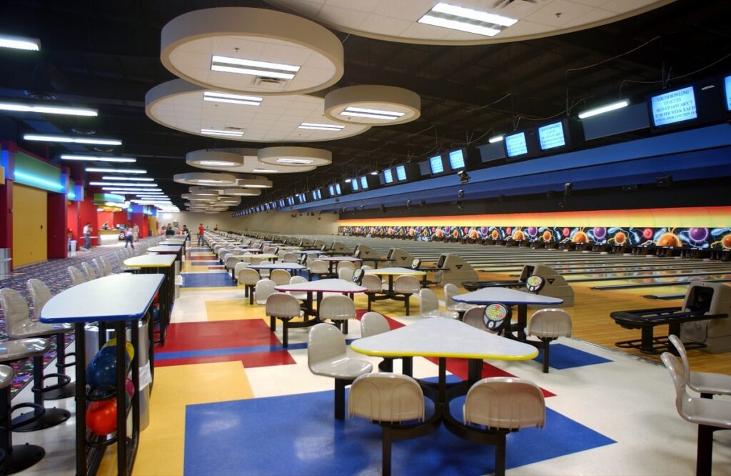 A brightly lit modern bowling alley featuring vibrant floors, neatly arranged tables and chairs, and several bowling lanes. Screens are mounted above each lane, with racks of colorful bowling balls lined up along the side.