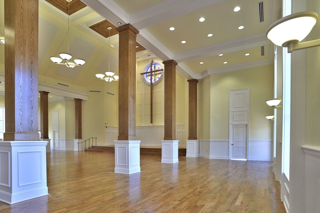 A large hall featuring wooden floors, tall columns, lofty ceilings with chandeliers, and a stained glass window above a modest stage. The space is illuminated by wall sconces and ceiling lights.