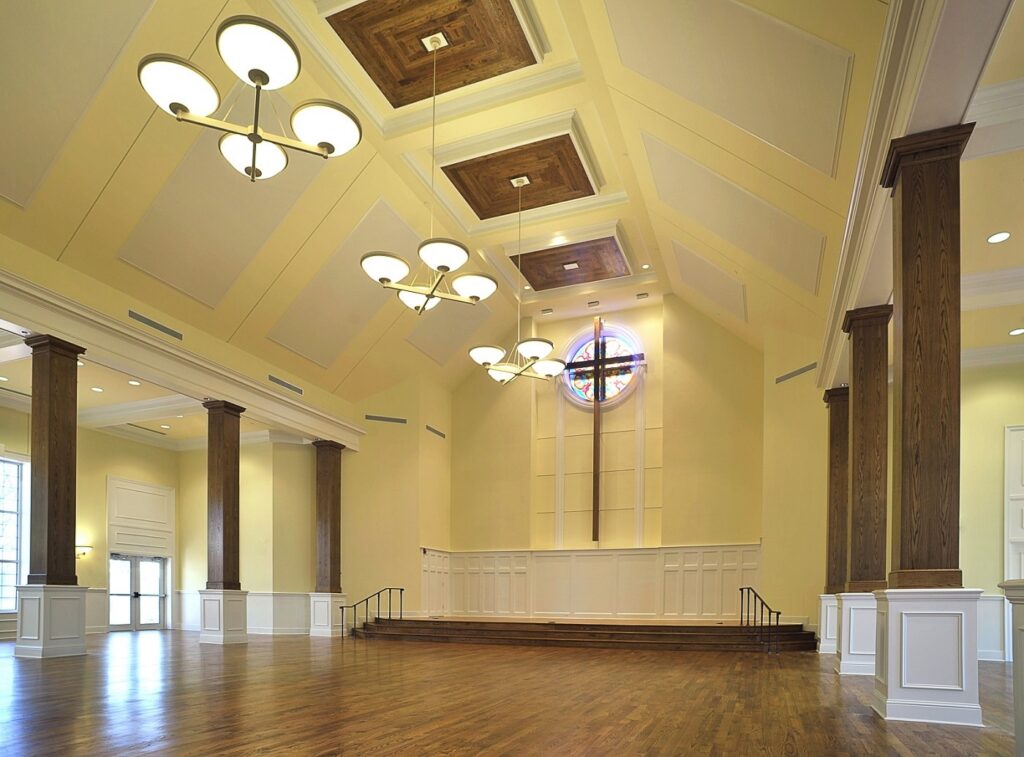 Spacious, elegant hall featuring wooden floors, tall pillars, a high ceiling with hanging lights, and a stage. At the far end is a cross beneath a stained glass window. The walls and ceiling are painted light yellow with white accents.
