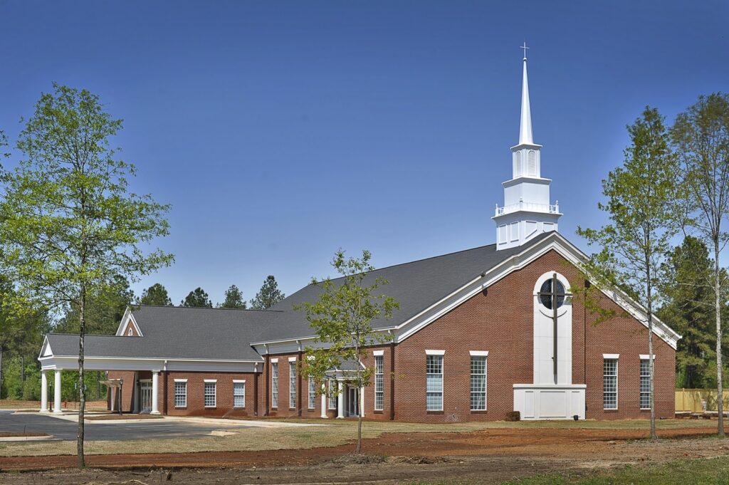 A spacious red-brick church featuring a tall white steeple, a large arched window, and several smaller windows, surrounded by young trees beneath a clear blue sky.