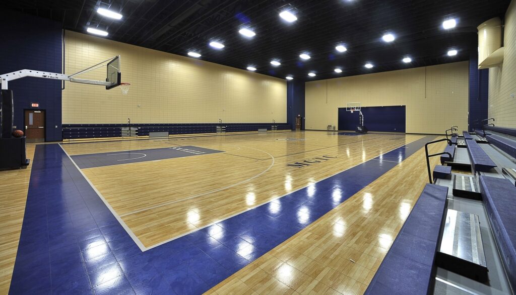 Empty indoor basketball court with wooden floor, blue and cream walls, two hoops, bleachers on the right, and bright overhead lighting. No people are present; the space is clean and well-lit.