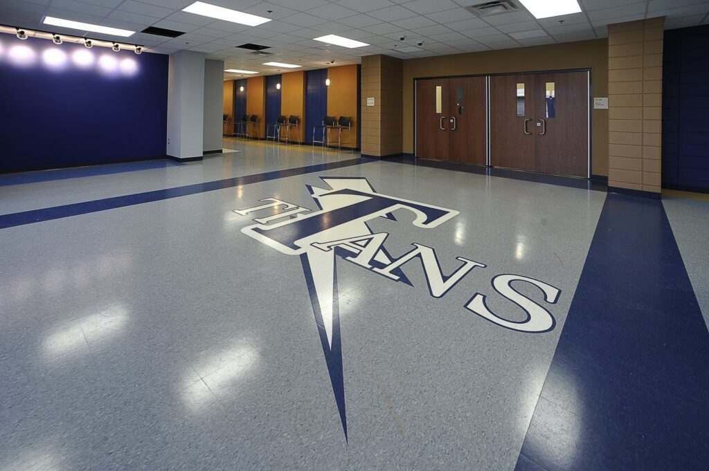 A school hallway with TITANS and a large T logo on the shiny floor, blue walls, and double wooden doors in the background.
