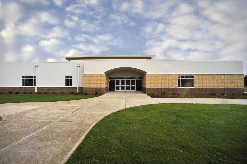 A contemporary single-story building featuring white and tan exterior walls, expansive windows, and a central glass doorway, set amidst a green lawn and bordered by a broad concrete walkway under a sky speckled with clouds.