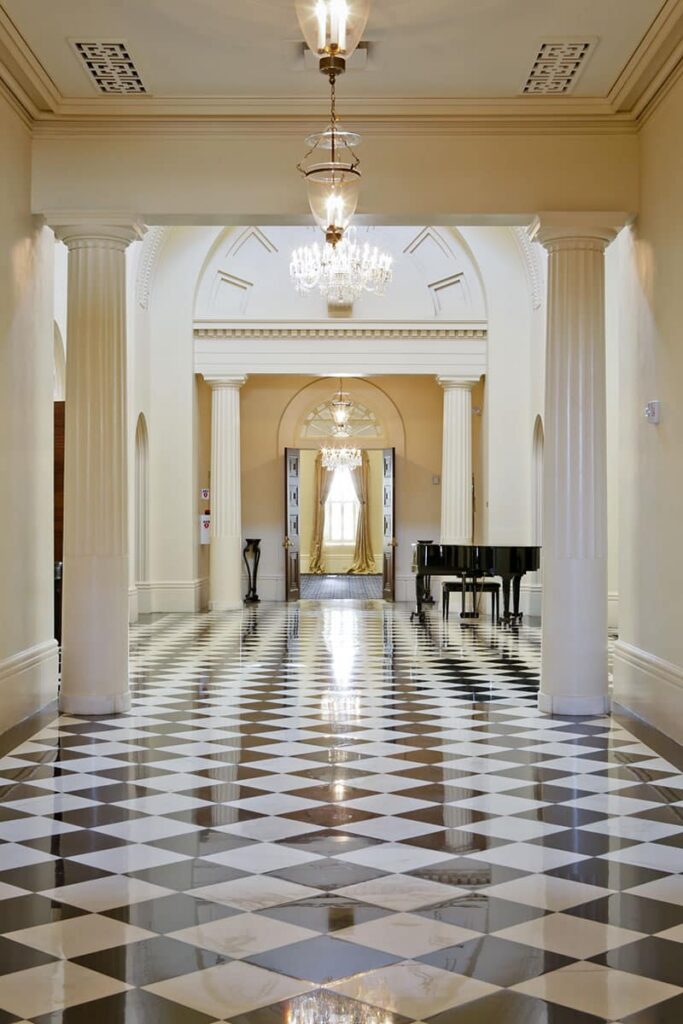 A spacious hallway features a black and white checkered floor, tall decorative columns, elegant chandeliers, a sleek black grand piano, arched doorways, and cream-colored walls that contribute to an overall atmosphere of elegance and luxury.