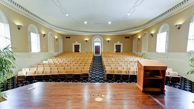 Looking out from a stage podium at an empty lecture hall featuring rows of beige seats, tall windows, arched doorways, and light-colored walls.