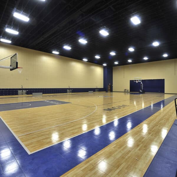 Empty indoor basketball court with wooden floors, blue painted lines, two basketball hoops, and blue bleachers along the right side. Overhead lights brightly illuminate the spacious gymnasium.