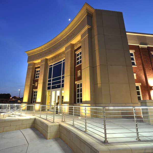 A modern, well-lit building made of brick and stone features large glass entrance doors and a curved facade. The photo is taken at dusk, capturing the deepening blue sky with a small moon visible above.
