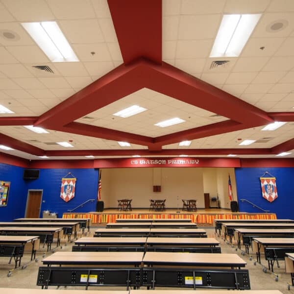 A well-lit school cafeteria features long tables, a stage at the front, colorful banners hanging on blue walls, and a ceiling decorated with bold red and white patterns.