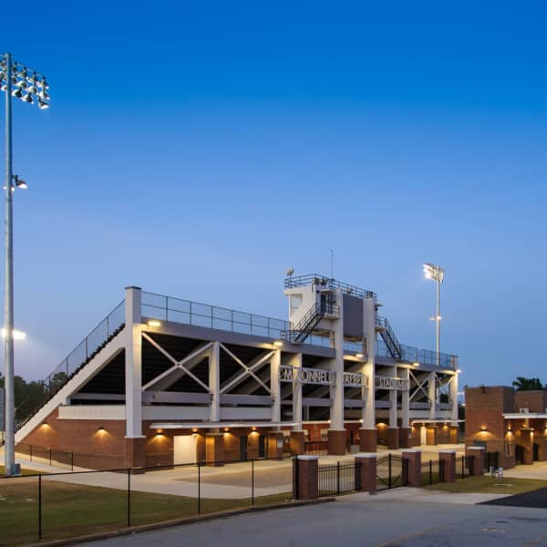 A football stadium illuminated by bright lights is empty at dusk. The modern building combines brick and metal elements, featuring a large central press box and several entryways beneath the bleachers.