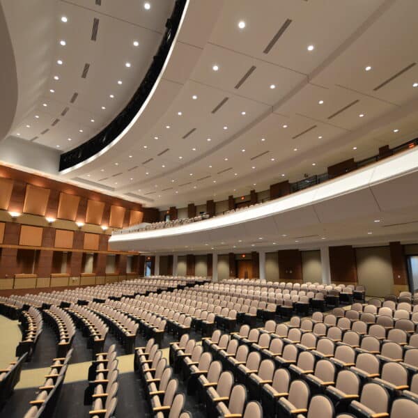 A spacious, empty auditorium featuring rows of beige seats, a curved stage on the left side, and an upper balcony. Recessed lights line the ceiling.