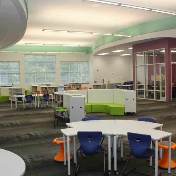 Modern library featuring white tables, blue and orange chairs, rows of bookshelves, and expansive windows. A purple accent wall and a glass-walled room appear in the background. The space is brightly lit, open, and arranged for collaborative learning.