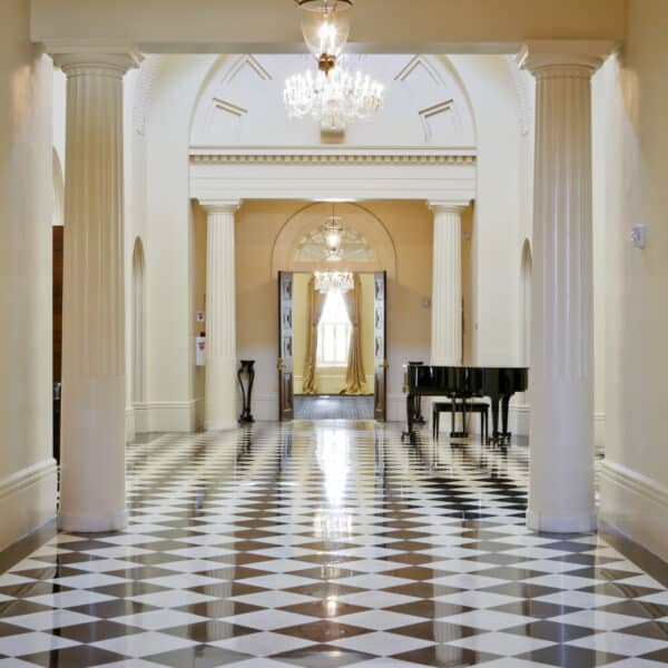 A spacious hallway features a black-and-white checkered floor, soaring columns, elegant chandeliers, creamy walls, and a black grand piano situated near the far end.